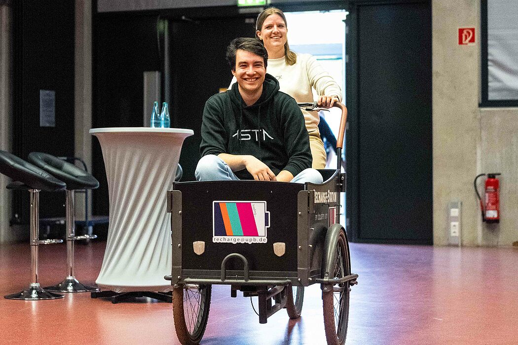Lena Wobbe, Mitarbeiterin des Hochschulsports, fährt mit dem Recharge-Fahrrad zur Erstsemesterbegrüßung. Im Fahrradanhänger sitzt Julian Rische, Vorsitzender des Allgemeinen Studierendenausschusses (AStA).