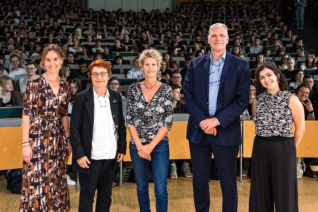 Im Vordergrund stehen Moderatorin Julia Ures, Universitätspräsidentin Prof. Dr. Birgitt Riegraf, Vizepräsident für Lehre, Studium und Qualitätsmanagement Prof. Dr.-Ing. Volker Schöppner, stellvertretende Bürgermeisterin der Stadt Paderborn Sabine Kramm und Sängerin Zara Akopyan. Im Hintergrund sitzen Studierende im Audimax.